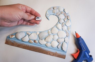 Hand placing a shell on a mosaic shell wave with a glue gun next to it