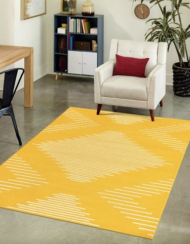 Bright yellow rug with white geometric shapes on a cement floor with the two front legs of an armchair resting on it.