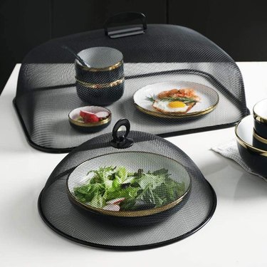 Stainless steel food covers in a black finish covering several plates of food. One cover is round and the other is rectangular.