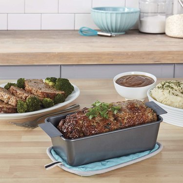 Finished meatloaf with herbal garnish shown in a Chicago Metallic meatloaf pan, pictured on a butcher block kitchen counter with gravy, potatoes and a plate of sliced  meatloaf behind it