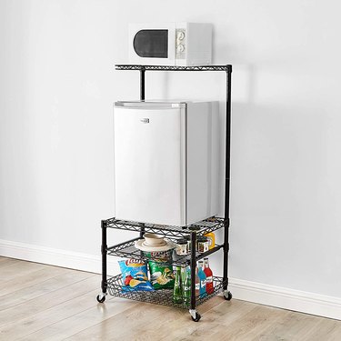 A rolling shelving unit with four shelves and cator wheels. A microwave sits on top and a mini fridge is on the second shelf.