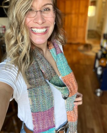 Smiling woman with blonde hair and glasses wears a white T-shirt and multicolored scarf