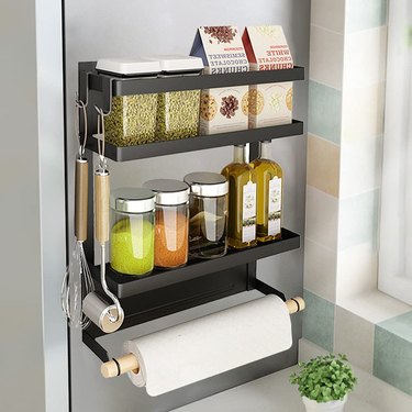 Magnetic spice rack with paper towel holder in black. There are hooks on the side for hanging utensils and a bar on the upper shelf prevents those items from falling.
