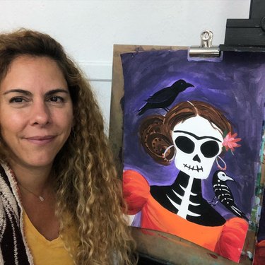 Woman with curly golden brown hair poses next to a Día de los Muertos painting