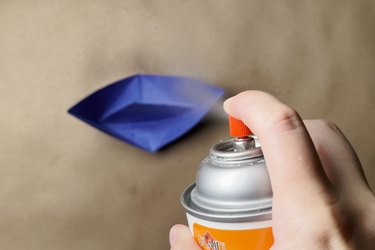 A spray can filled with clear acrylic sealer spraying a blue paper boat