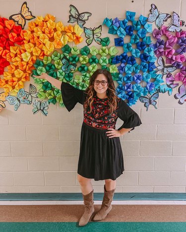 Caucasian woman wearing a black dress with a floral print points to a wall of rainbow-colored paper art