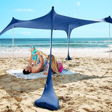 Navy blue canopy tent on a beach with parents and a baby laying down on a blanket under the canopy.
