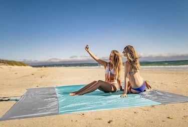 Two girls at the beach taking a selfie on a Wekapo Sandproof Beach Blanket in blue and gray.