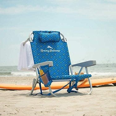 Tommy Bahama Beach Chair pictured on the sand at the beach. The chair has a blue foral design, a padded headrest, cup holder, and a bar for hanging your towel.