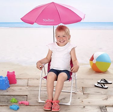 Kid's Beach Chair and Personalized Umbrella Set pictured here in pink. A little girl with blone hair is sitting on the chair and smiling. There is a fake beach background and beach toys scattered about.
