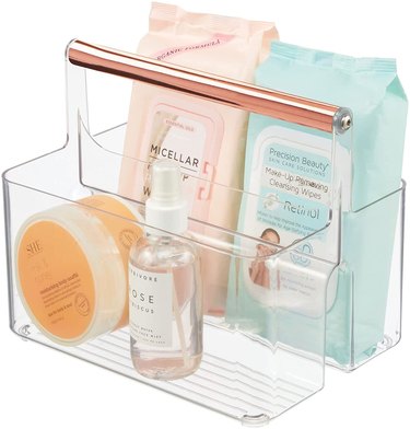 Clear shower caddy with rose gold handle and shower items inside.