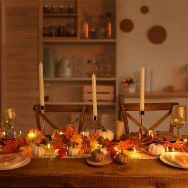 Fall table set for guests with the lights down low. There are three tall candlesticks in the center and a leaf garland with LED lights in it running down the center of the table.