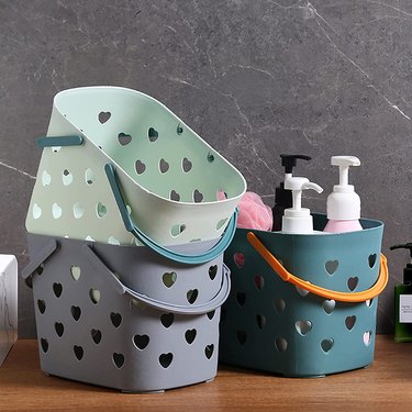 Three plastic shower caddies with heart-shaped drainage holes on a wooden table surrounded by gray marble.