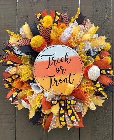 Candy Corn Wreath from Etsy. The center is a candy corn-colored circle that says "Trick or Treat" in cursive lettering and there are rolls of different colored fabric and ribbon surrounding it.