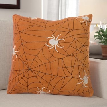 Orange Velvet Throw Pillow from Wayfair with a Black Spider Web Design and Multiple White Spiders Embroidered Onto It.