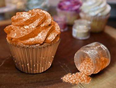 Pumpkin Orange Tinker Dust Edible Glitter from Etsy in a 5-gram jar, sprinkled on a cupcake's frosting in a metallic wrapper.
