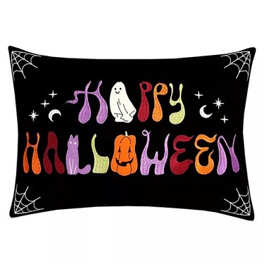 Happy Halloween Throw Pillow from Kohl's. The pillow is black with colorful letters that spell "Happy Halloween." The A is a ghost and the O is a pumpkin, both are grinning.