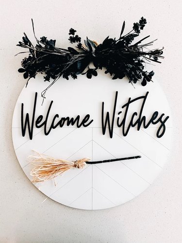 Welcome Witches Halloween Front Door Sign from Etsy. It's circular and painted white with a herringbone-like design etched into the wood. The script writing is black, there's a decorative broom on the bottom, and black faux florals at the top.