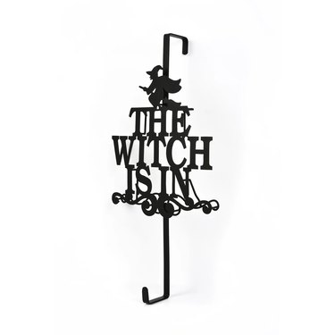 Wreath Hanger from Wayfair that says "The Witch Is In" with the image of a witch flying on a broom. It hooks over the top of your door, so no nail is necessary.