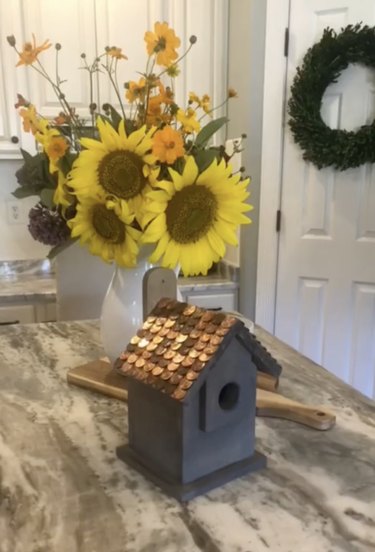 Wooden birdhouse with penny roof sitting next to bouquet of yellow sunflowers
