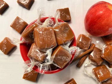 Pile of brown caramels topped with salt arranged in a small red bowl alongside a red apple