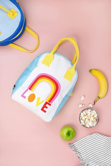 Blue, yellow, and white lunch tote with a rainbow and the word "LOVE" underneath it against a pink background with snacks and the corner of a second lunch tote.