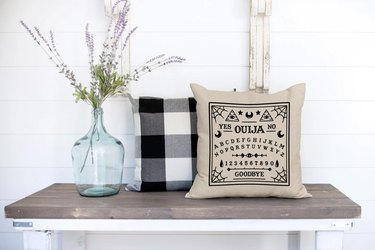 Tan throw pillow with ouija board design on it in black lettering on a bench next to a black and white buffalo check pillow.