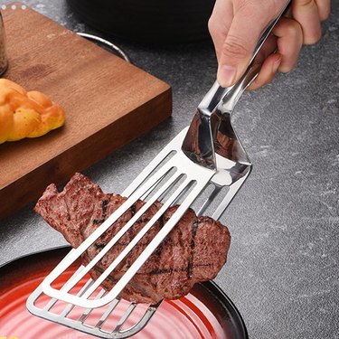 Hemoton Stainless Steel Fish Spatula being used to pick up a piece of meat. It looks like a slotted smaller spatula and a larger slotted spatula connected to act like tongs.