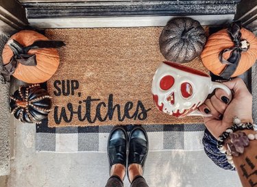 "Sup Witches" doormat with decorate pumpkins and person holding a spooky mug