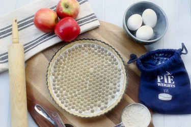 Overhead view of a tart pan lined with crust, parchment and Gretel brand glass pie weights, with extra weights at the side in their drawstring bag