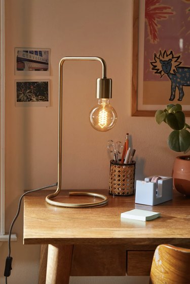 Brass-finish desk lamp with a circular wire base and an exposed Edison bulb on a wooden desk.