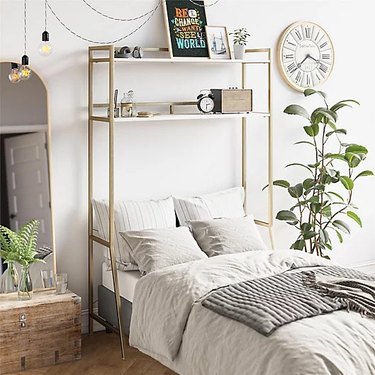 An over-the-bed shelf in gold and white that fits over a Twin XL bed. On the shelves are framed artwork, an alarm clock, headphones and a speaker.