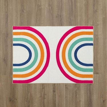 White rectangular rug with two multi-color rainbow arches on either side (left and right) against a wood floor.