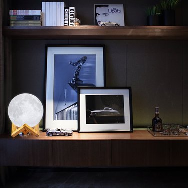 Moon lamp in a dimly lit room sitting on the included wooden stand next to framed photos and a car figurine.