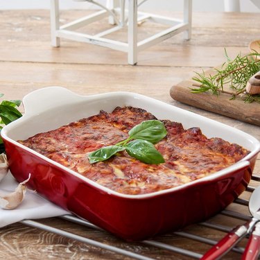 Lasagna baked in a red Emile Henry casserole dish, garnished with fresh basil, depicted on a metal trivet on a wooden countertop