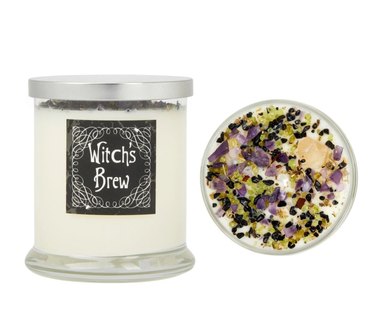 White candle in glass jar reading "Witch's Brew" topped with black, purple and green stones