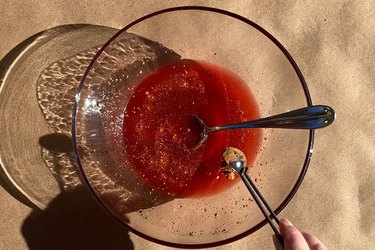 A hand adding cocoa powder to a clear bowl filled with water, corn syrup and red food coloring