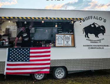 Alex stands at the counter of his white food truck that sports a buffalo logo next to the counter, an American flag below and string lights above.