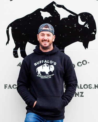 Alex leans in front of the truck's buffalo logo, smiling, wearing a backwards cap, and a black Buffalo's American Food Co. hoodie.