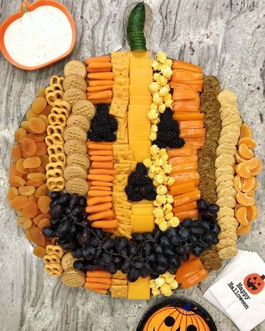 oranges, pretzels, crackers, cheese, popcorn, berries, cookies and grapes arranged to form a pumpkin face