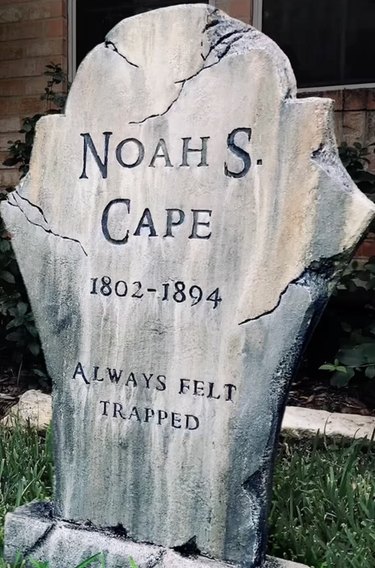 Fake tombstone reading "NOAH S. CAPE, 1802-1894, ALWAYS FELT TRAPPED"