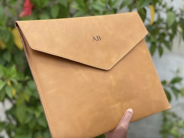 Light brown leather laptop sleeve with initials in an anonymous person's hand against a background of greenery.