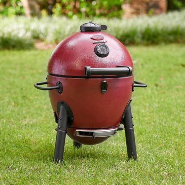 Red Akorn Jr. kamado grill, shown on a grassy lawn with a flower bed in blurred focus in the background