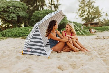 Two women sitting on a beach underneath a striped umbrella tent that's staked down to the ground.
