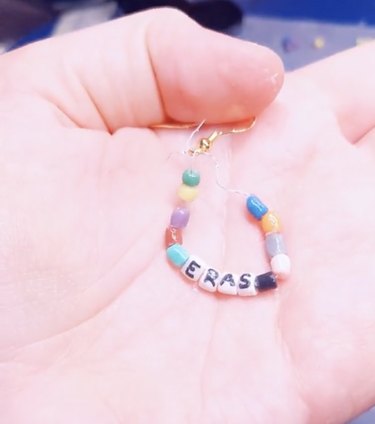 Small beaded earrings reading "ERAS" sitting on a light-skinned hand