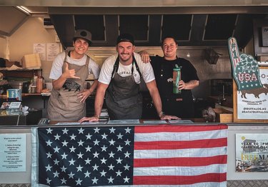 Alex Moore and his team pose in the window of their food truck, decorated with an American flag.