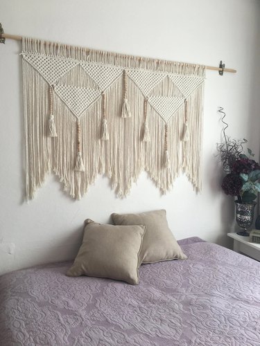 Macrame wall hanging on a wooden dowel with tassels hanging behind a bed with a lilac quilt and two tan throw pillows.