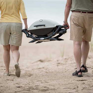Two anonymous people carrying a Weber Q2200 on a sandy beach.