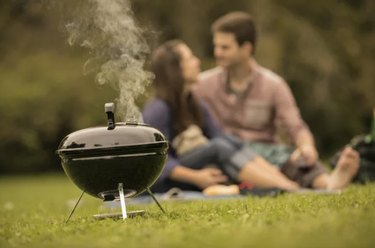 A small Weber Smokey Joe on the grass, releasing a bit of smoke. Two people are sitting on a picnic blanket in the background.