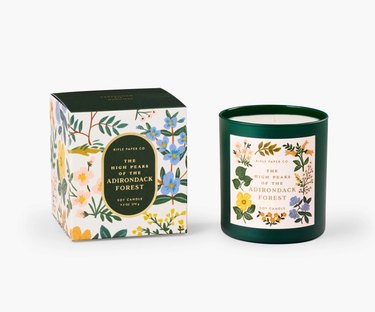 Rifle Paper Co. High Peaks of the Adirondack Forest Candle next to its decorative box. The glass candle jar is dark green and the label has a floral design on it.